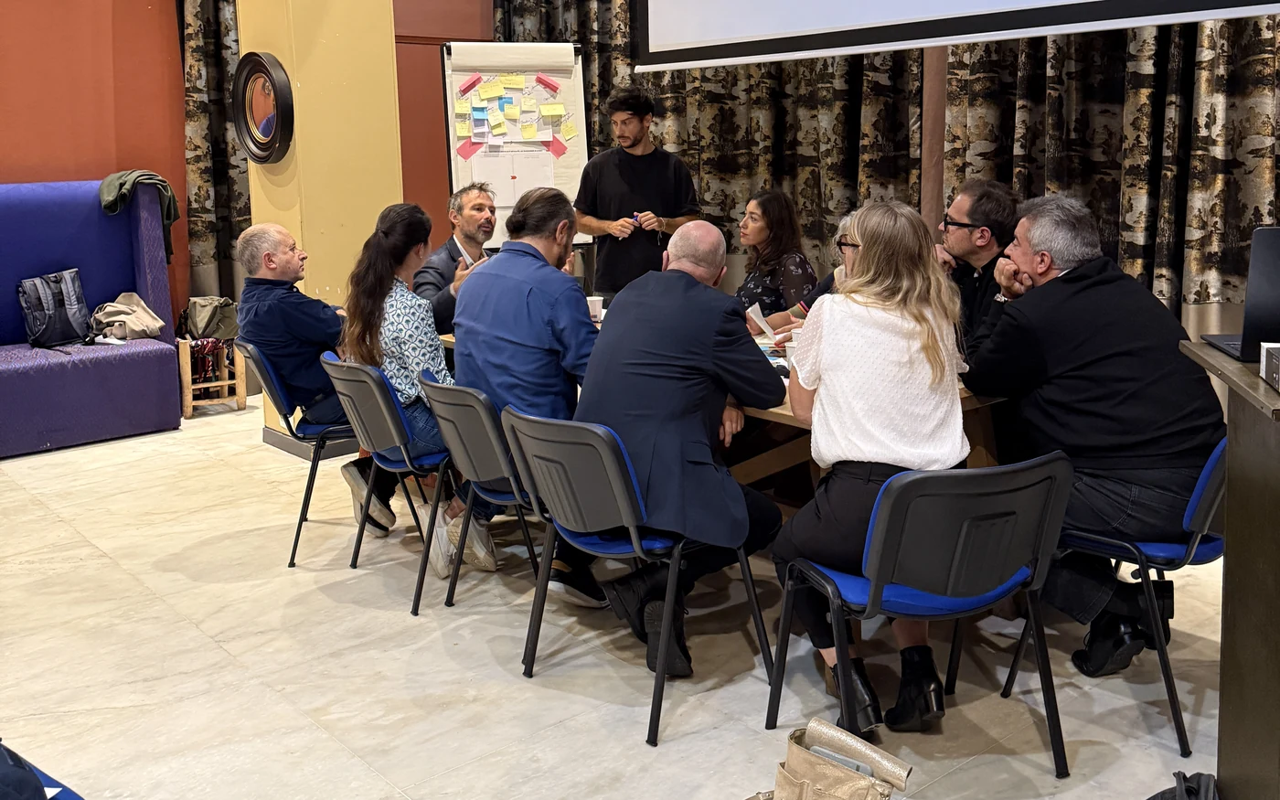 Une équipe assise autour d'une table de travail, un animateur debout devant un paperboard de post-it